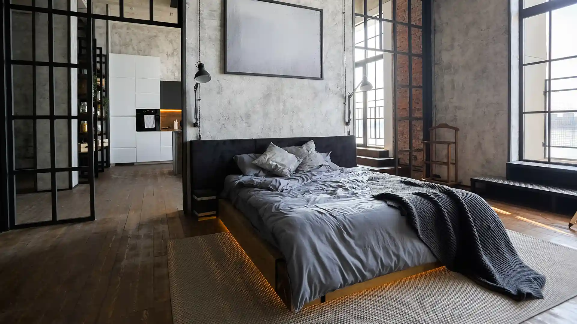 Ein ungemachtes Bett steht in einem industriellen Schlafzimmer mit großen Fenstern, flankiert von moderner Beleuchtung. Im Hintergrund befindet sich eine offene Küche mit weißen Schränken und Holzfußboden.
