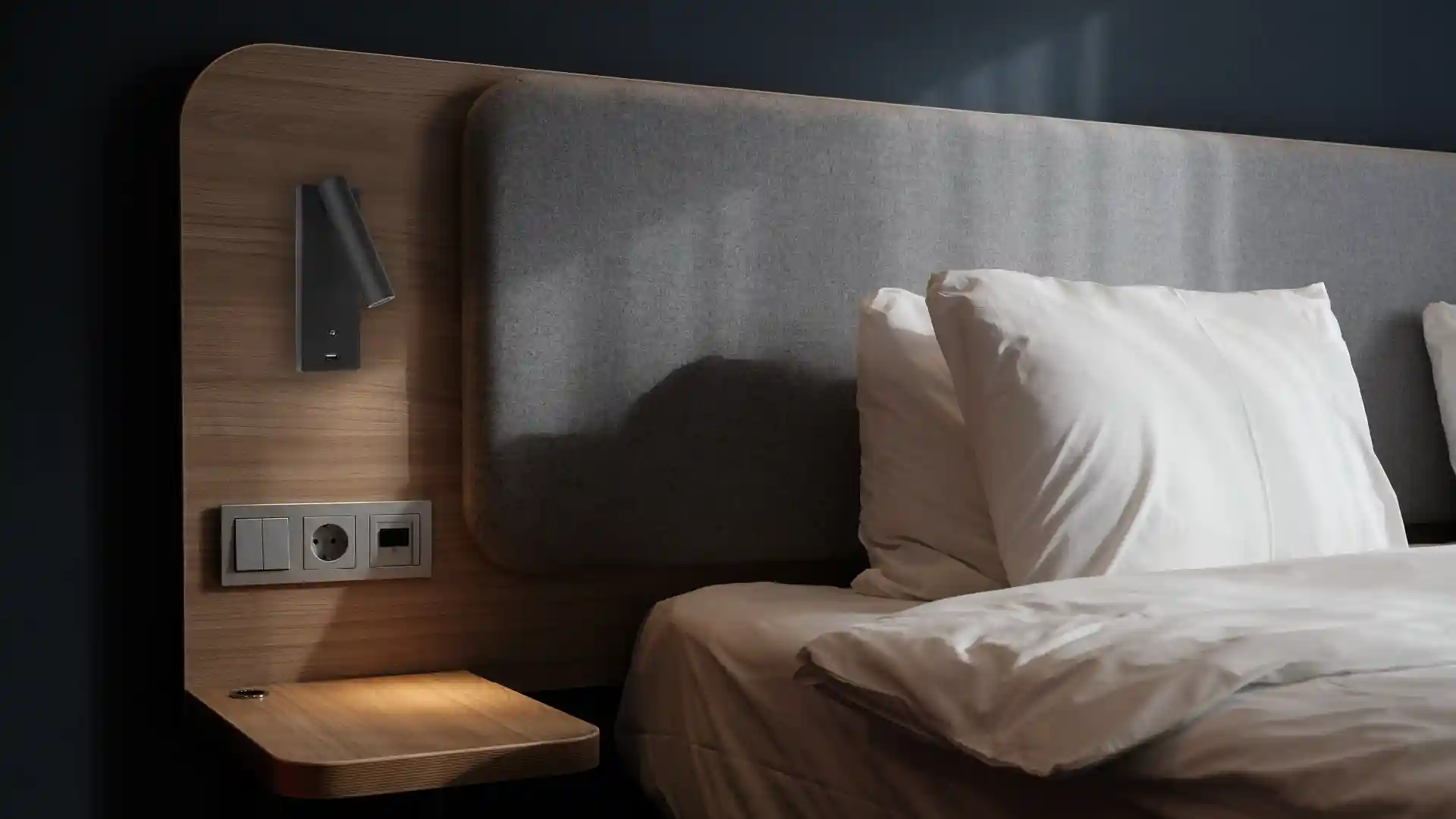 Ein Bett mit weißen Kissen steht neben einem Holz-Nachttisch mit Steckdosen, Lichtschalter und einer kleinen Lampe; die Wand ist in einem dunklen Ton gehalten.