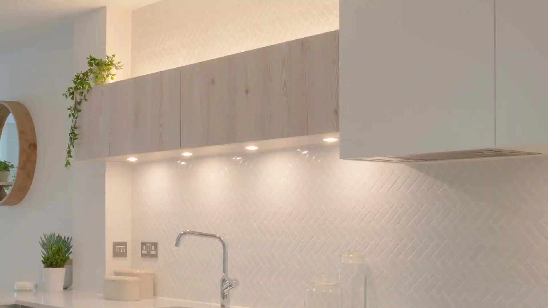 Ein moderner Wasserhahn steht über einem weißen Spülbecken auf einer glänzenden Küchenarbeitsplatte, umgeben von eleganter Beleuchtung, einem Weinglas und Geschirr vor einem minimalistischen, hellen Hintergrund.