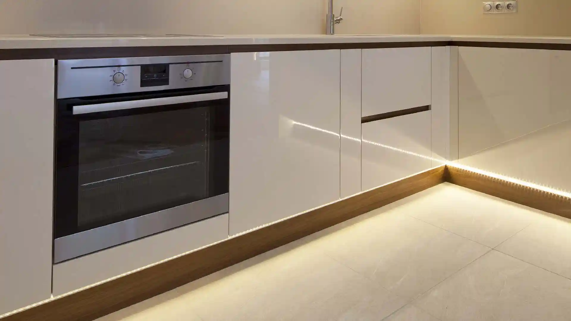Ein moderner eingebauter Ofen mit silbernem Bedienfeld ist in einem weißen, glänzenden Küchenschrank integriert. Weiche Beleuchtung betont den Boden und die Ecke der minimalistischen Küche.