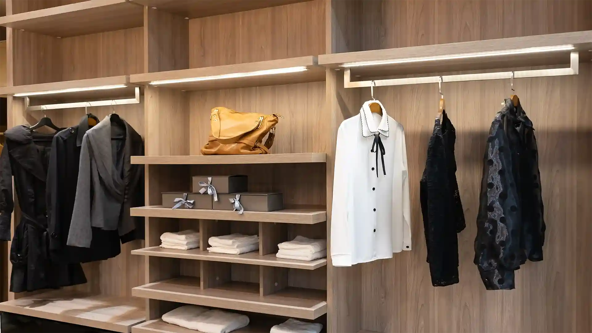 Ein Kleiderschrank aus Holz mit hängenden Hemden und Mänteln auf der linken Seite, einem braunen Beutel auf dem Regal, und gefalteten Handtüchern sowie Geschenkboxen in der Mitte.