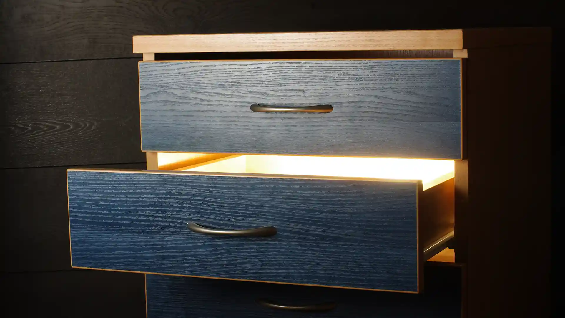 Ein Holztisch mit offenen, leuchtenden Schubladen in blauem Finish vor einer dunklen Wand.