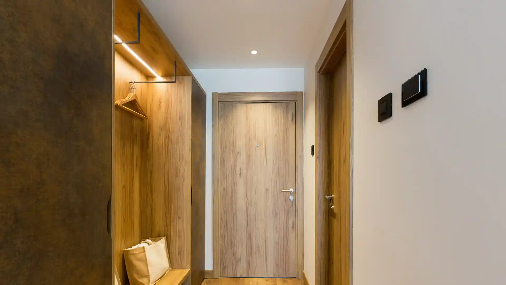 Eine Holztür führt in einen schmalen Korridor mit Holzinterieur; links eine beleuchtete Garderobe mit Kleiderbügeln und Tasche, rechts ein weißer Lichtschalter an der Wand.