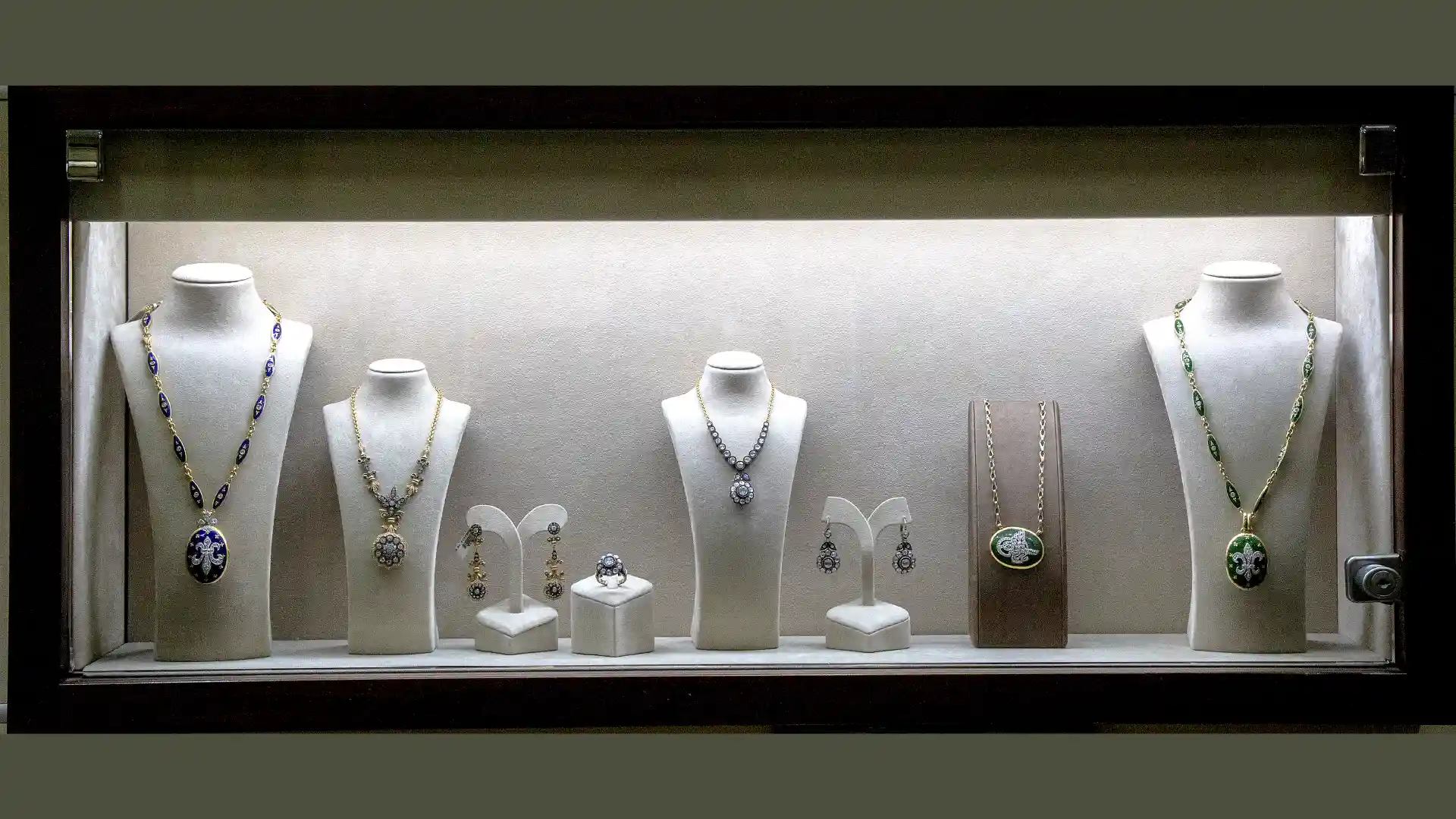 Eine Sammlung eleganter Schmuckstücke, darunter Halsketten, Ringe und Ohrringe, wird auf weißen Büsten und Ständern in einer beleuchteten Vitrine ausgestellt.
