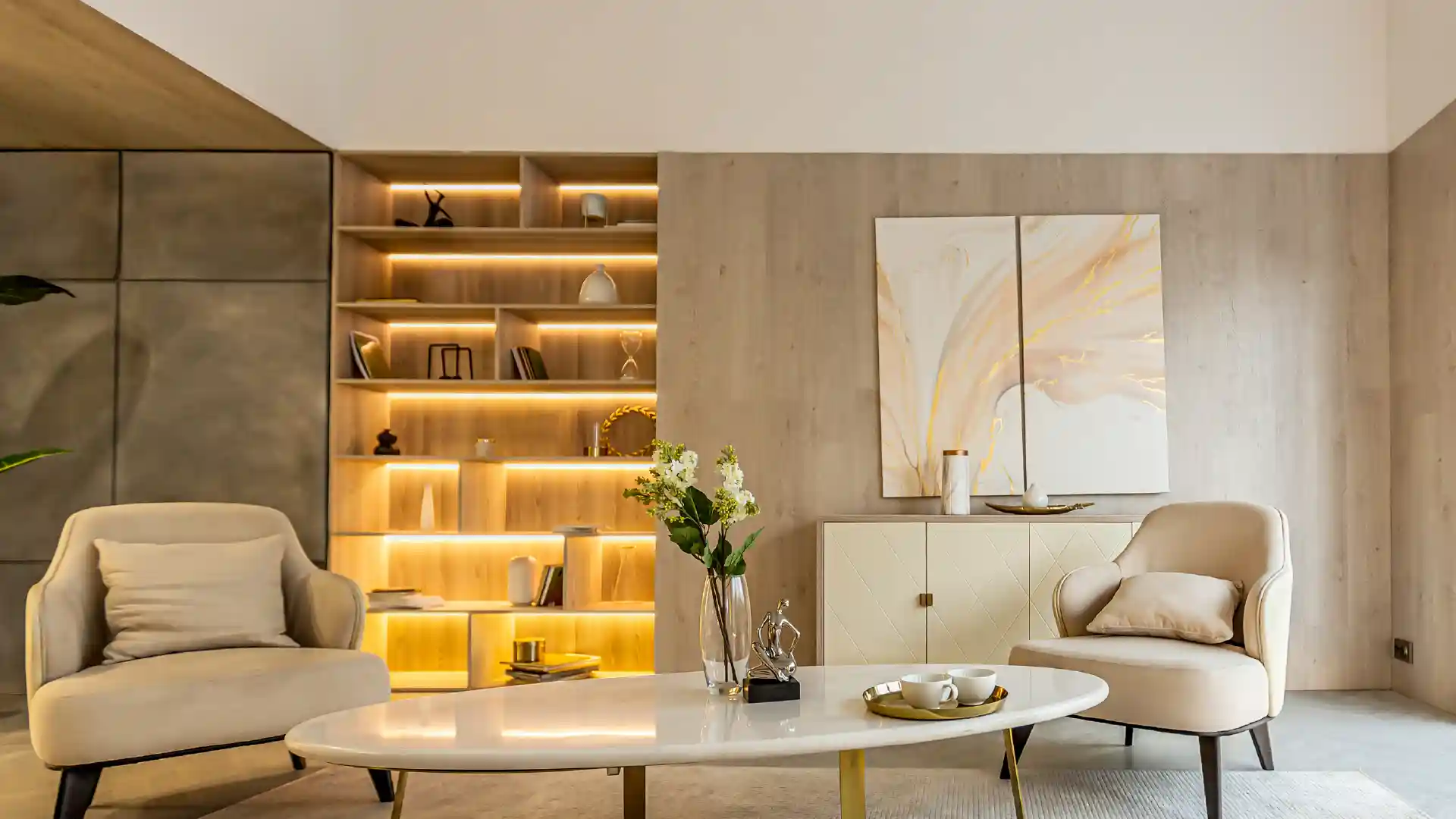 Ein Wohnzimmer mit zwei beigen Sesseln, einem Couchtisch, auf dem Blumen und Tassen stehen. Im Hintergrund ein beleuchtetes Bücherregal und eine zweigeteilte Kunstwand.